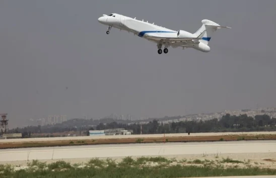 A white and blue surveillance aircraft, the focus keyphrase of this scene, takes off from a runway under a cloudy sky, with a cityscape and trees visible in the background.