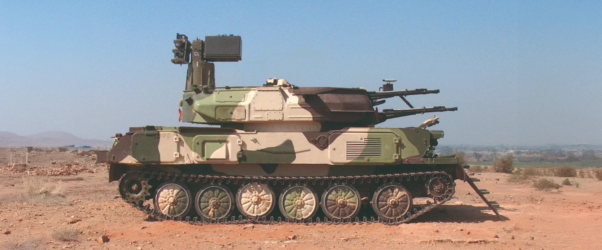 A camouflaged military tank with twin cannons and radar equipment on top, designed for mobile air defence, is parked on dry, sandy ground with hills and sparse vegetation in the background.