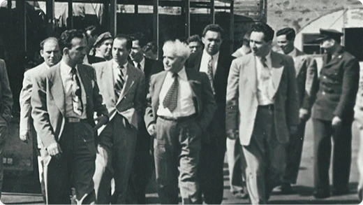 A group of men in suits walk together outdoors in a black-and-white photo; a few soldiers or officials stand in the background near a building or vehicle.