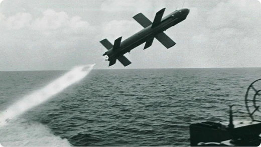 A black and white photo of a missile being launched over the ocean, leaving a trail of smoke behind, with part of a ship visible in the bottom right corner.
