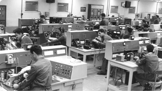 Black and white photo of a busy electronics workshop, with several men in work coats seated at benches, assembling or testing electronic equipment surrounded by various technical instruments.