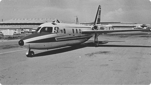 Black and white photo of a small jet airplane parked on a runway, with a large hangar and other airport structures visible in the background.