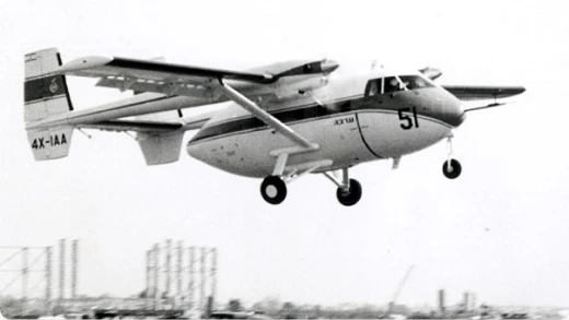 A twin-engine, high-wing aircraft with the number 51 on its nose is flying low above an industrial area. The registration 4X-IAA is visible on the tail. The image is in black and white.