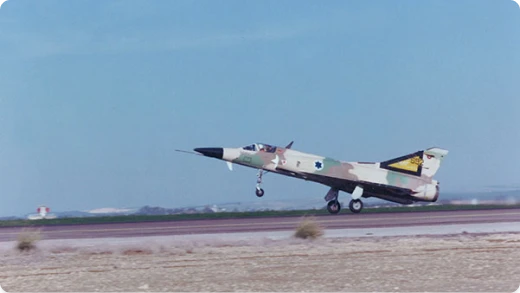 A military jet with desert camouflage and an Israeli Air Force insignia takes off from a runway against a clear blue sky.