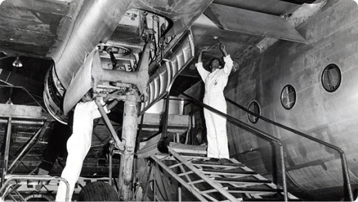 Two workers in white coveralls stand on a staircase, inspecting and working on the underside of a large aircraft inside a hangar. The aircraft’s round windows are visible in the background.
