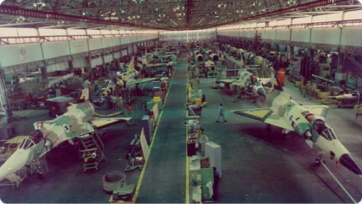 A large aircraft assembly plant with several jet fighters under construction, workers and equipment spread throughout the spacious, high-ceilinged facility, and bright lighting overhead.