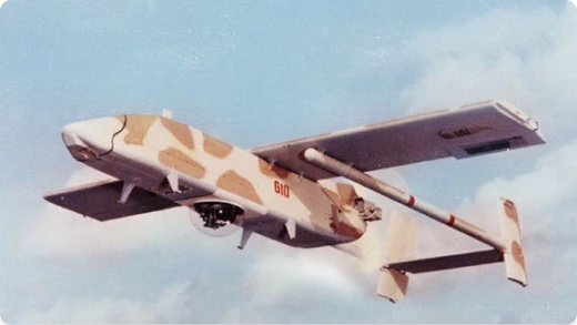 An unmanned aerial vehicle (UAV) with tan and brown camouflage flies against a partly cloudy sky. The drone has a boxy fuselage, straight wings, and a rear-mounted propeller. The number 610 is visible on its side.