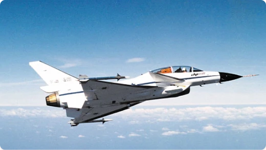A white and gray jet fighter aircraft with a tandem cockpit is flying above the clouds against a clear blue sky.