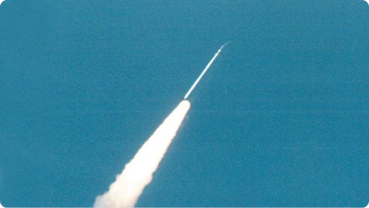 A rocket ascends into a clear blue sky, leaving a thick white trail of smoke behind it.