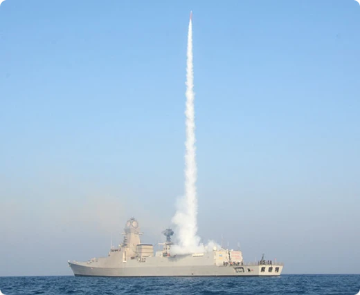 A warship at sea launches a missile vertically, creating a tall white smoke trail against a clear blue sky.