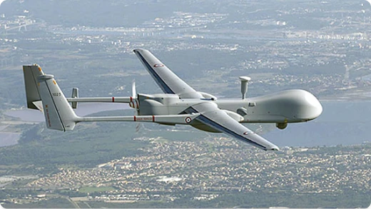 A large military drone flies above a city and landscape, featuring a long wingspan, twin tail booms, and a distinctive rounded nose, with visible markings on its body.