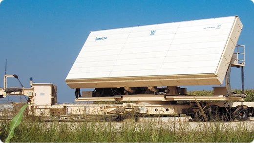 Large white military radar system mounted on a beige trailer sits on grassy terrain under a clear blue sky.