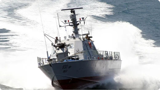 A military patrol boat speeds through the ocean, creating a large wake behind it. Several crew members are visible on deck, and the vessel is marked with the number 828.