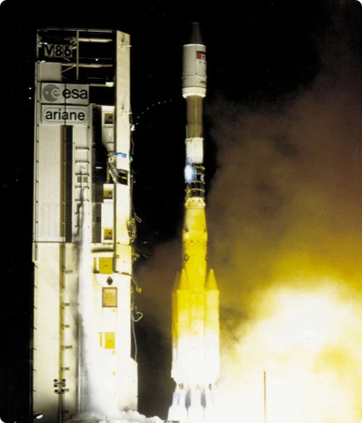A rocket labeled ESA ariane launches at night, emitting bright yellow flames and smoke from its base. The scene is dark, with the rocket and launch structure illuminated by the launch light.