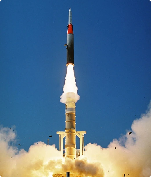 A rocket launches vertically into a clear blue sky, emitting bright flames and smoke from its engines at the base, with debris scattered around the launch pad.