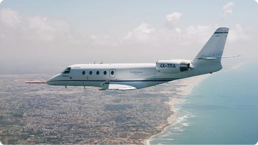 A white private jet with the registration 4X-TRA flies over a coastal city, with the ocean on one side and urban buildings below, under a partly cloudy sky.
