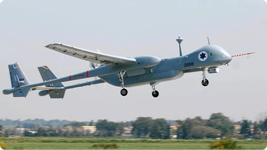 A gray military drone with a blue Star of David emblem on the side is flying above a grassy field, with trees and buildings blurred in the distant background.