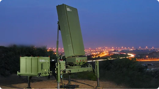 A green military radar system is set up on a hill at dusk, with a vertical rectangular radar panel and equipment box. City lights and a winding road are visible in the background.
