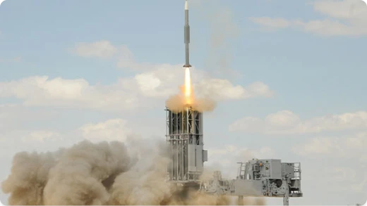 A missile launches vertically from a ground platform, producing a trail of smoke and fire against a partly cloudy sky.