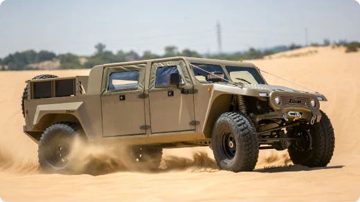 A rugged, tan off-road vehicle with large tires drives through sandy terrain, kicking up dust. The vehicle has four doors and an open cargo bed, with a desert landscape in the background.
