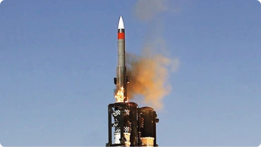 A missile is launching vertically into a clear blue sky, with flames and smoke visible at its base as it rises from a multi-barrel launcher.