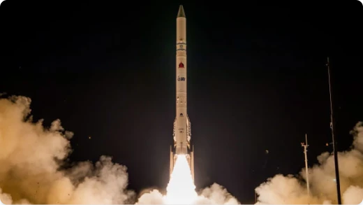 A white rocket launches vertically into the night sky, surrounded by bright flames and clouds of smoke at its base. The rocket is illuminated against a dark background.