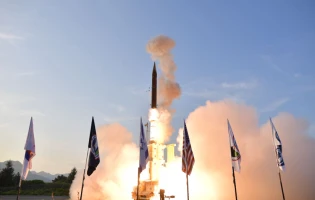 A missile is being launched into the sky with clouds of smoke at the base during the Le Bourget Paris Air Show. Several flags are displayed in the foreground, and the scene is set outdoors under a clear blue sky.