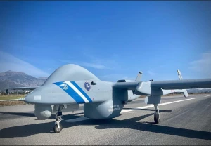 A large gray military drone with blue and white stripes is positioned on a runway under a clear blue sky, with mountains visible in the background.