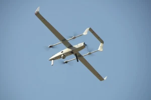 A white drone with a unique U-shaped tail design and four propellers soars beneath the clear blue sky, resembling a mini satellite in flight.