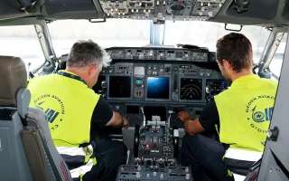 Two aviation technicians wearing yellow vests sit inside an airplane cockpit, facing the instrument panel as they perform maintenance or system checks.