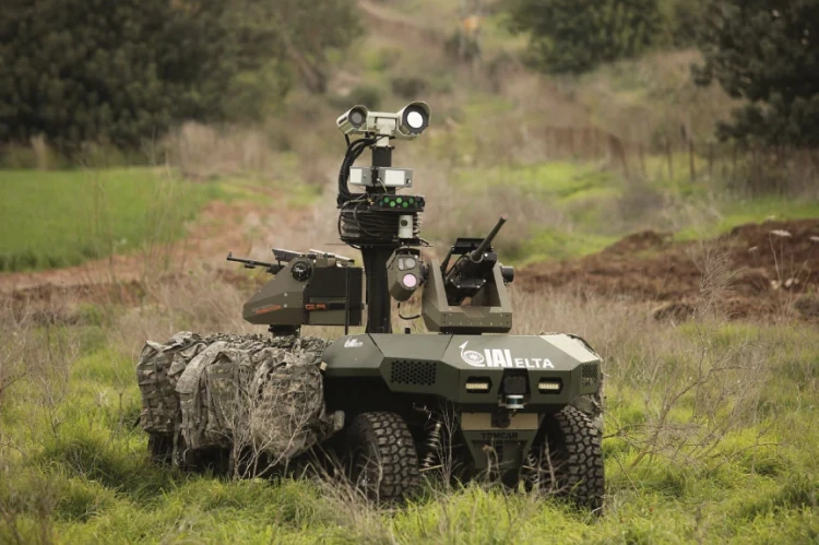 A military ground robot, part of modern warfare systems, is equipped with cameras, sensors, and mounted weapons. It stands on grassy terrain, surrounded by greenery and trees in the background.