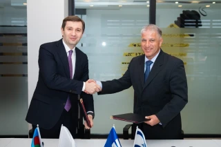 Two men in suits shake hands across a table with documents, IAI flags displayed, appearing to finalize an agreement or partnership in a modern office setting.