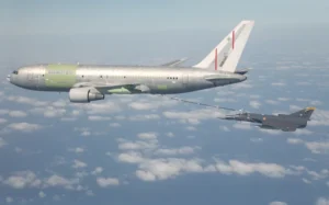 An IAI military tanker aircraft refuels a fighter jet mid-air over the ocean, both flying above scattered clouds. The tanker has an unpainted, metallic appearance with red and green markings.