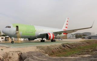 A large commercial airplane with a green and white fuselage and red-and-white striped tail is parked near an industrial area, where Air defence Systems are visible in the background alongside a work ladder at the front.