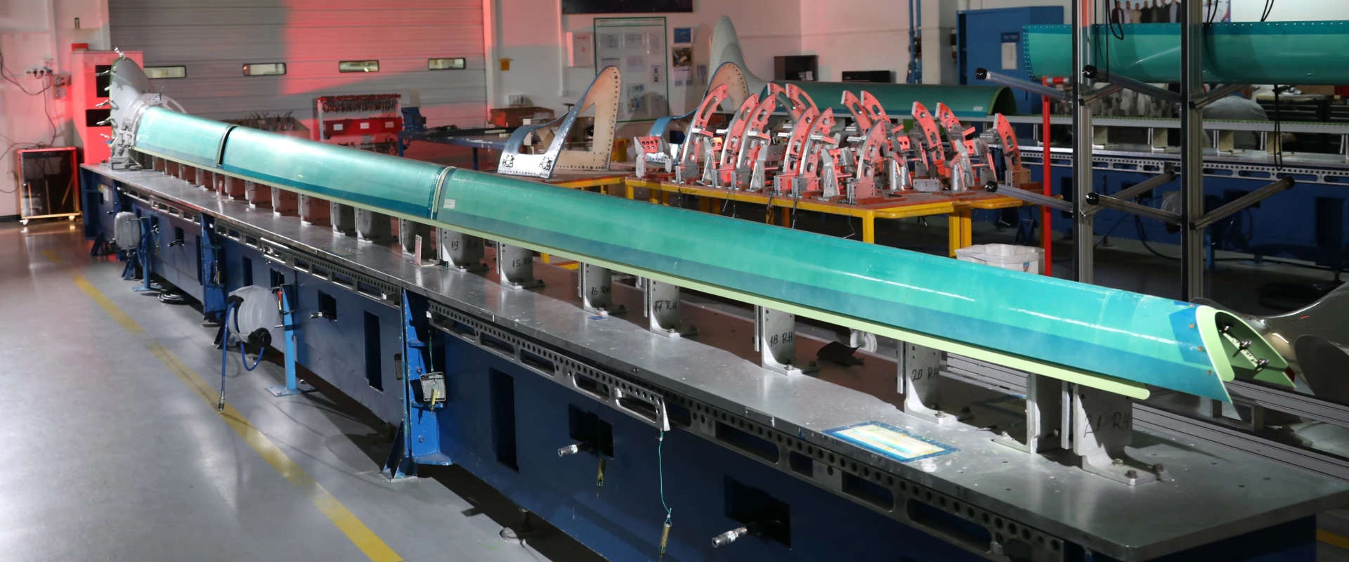 A large, green composite aircraft wing panel, part of the B777/B777X Empennage Assemblies, is positioned on a metal assembly table in a brightly lit industrial workshop with various tools, machinery, and equipment in the background.