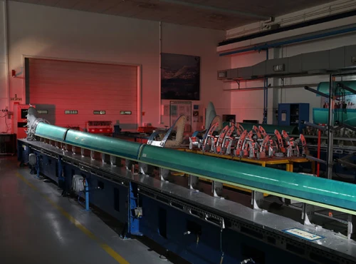 A long, green aircraft wing is being assembled in a factory, surrounded by tools and industrial equipment under dim lighting—highlighting the precision involved in B777/B777X Empennage Assemblies.