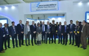 A group of men and women in formal attire, including military officers in uniform, stand posing for a group photo in front of a Bharat Electronics exhibition booth at an indoor event.