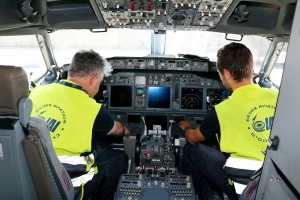 Two people wearing yellow vests are seated in an airplane cockpit, operating controls and instruments as they perform commercial aviation maintenance inside the aircraft.