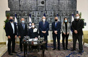 A group of seven people, all wearing face masks, stand and sit in front of Israeli flags and a large wall sculpture. A small round table with items on it is in front of them.