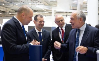 Four men in suits are standing together indoors, engaged in conversation. One man gestures with his hand while the others listen and smile. The background shows a modern, well-lit industrial or conference setting.