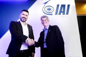 Two people in suits smile and shake hands in front of a white wall with the blue IAI logo, which includes a stylized star and swoosh symbol, during a cysource partnership event in Israel.