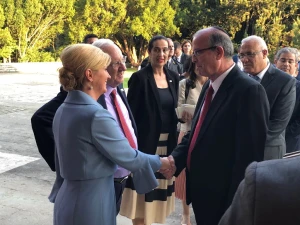 A woman in a light blue outfit, focused on contamination control, shakes hands with a man in a black suit and red tie outdoors as several people watch the greeting amid trees and greenery.