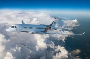 A sleek white Dassault Aviation Israel Falcon 10X private jet flies above the clouds, with a blue sky and distant land below. The jet has red and dark accents, sunlight illuminating its fuselage and wings.