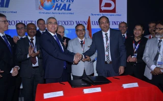 A group of men in suits stand behind a table with red cloth as two men in the center shake hands, smiling—finalizing a deal. Papers and pens are on the table, and company logos highlighting potential market share are displayed on a screen in the background.