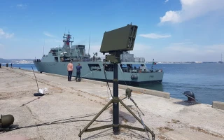 A military ship is docked at a pier, with a tripod-mounted radar or communication device set up in the foreground. A few people stand near the ship. The sky is partly cloudy.