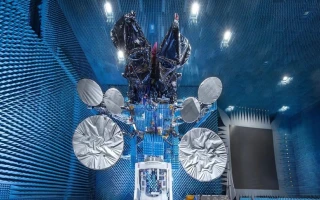 A satellite with large, silver circular antennas is suspended in an anechoic chamber, resembling a scene from the IAI annual report, surrounded by blue, sound-absorbing foam panels and bright overhead lights.