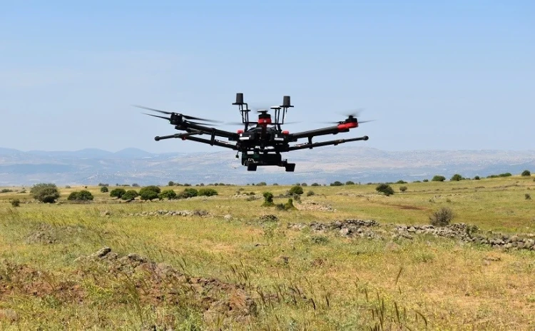 A large black IAI ELTA drone with multiple propellers flies over a grassy open field, likely equipped with SAR technology, amid scattered bushes and distant hills under a clear blue sky.