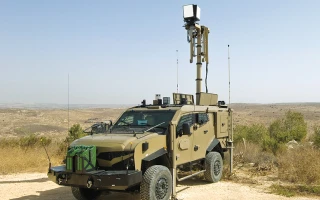 A military armored vehicle equipped with advanced surveillance equipment and sensors mounted on a tall mast, parked on a dirt road in a dry, open landscape.