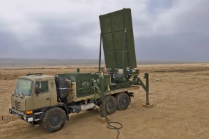 A military truck with a large, upright radar system is positioned in a desert landscape under a cloudy sky. The radar dish is elevated and attached to the truck’s rear section.