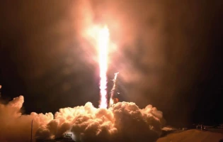 A rocket launches into the night sky, leaving a bright trail of flames as it carries the EROS C3 satellite into space. Thick clouds of smoke billow around the launch pad against a dark background.